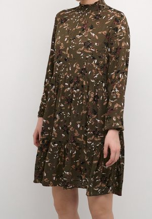 Femme portant une robe fleurie vert olive avec manches longues, un col montant froncé et une jupe à volants sur un fond clair uni.
