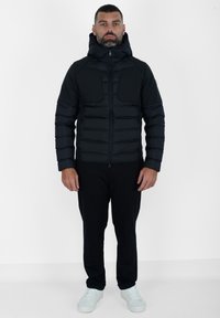 Giacca a vento nera con cappuccio, caratterizzata da una texture trapuntata, due tasche frontali e chiusura a zip. Indossata con pantaloni neri e sneakers chiare.
