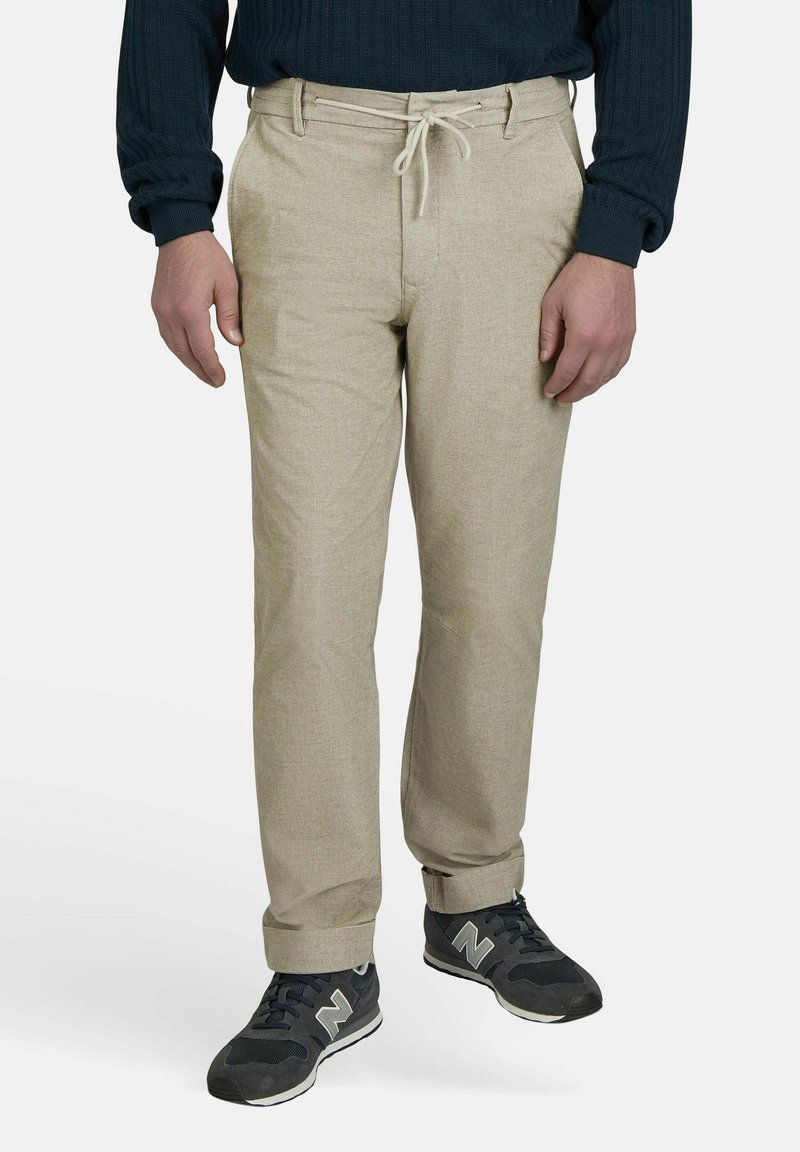 Beige broek met een textuurafwerking, voorzien van een trekkoord in de taille. Gecombineerd met donkergrijze sneakers met witte accenten.