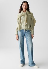 Lichtgroene blouse met lange mouwen en een trekkoord in de taille, gecombineerd met wijduitlopende vervaagde blauwe jeans en een gestructureerde lichtgroene wikkel. Witte schoenen.