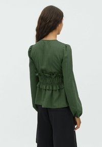 Green, textured blouse with smocked waist and ruffled detail. Long, puffed sleeves. Styled with black, tailored shorts.
