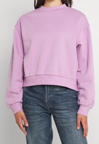 Fioletowy, skrócony sweatshirt z wysokim kołnierzem, ściągaczami oraz miękką fakturą, w zestawieniu z ciemnoniebieskimi dżinsami.