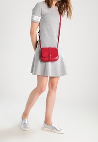 Röd läderaxelväska med en platt rektangulär form, ett lockstängning och en lång rem. Bärs med en grå klänning och silverfärgade slip-on skor.