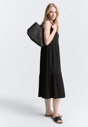 Robe noire sans manches avec une jupe à volants, associée à un sac fourre-tout noir texturé et des sandales noires à glisser, sur un fond clair.