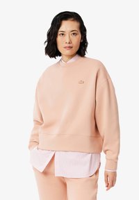 Rosa hettegenser laget av mykt materiale, har rund hals, ribbestrikkede mansjetter og en liten logo. Laget over en rosa og hvit stripet skjorte.