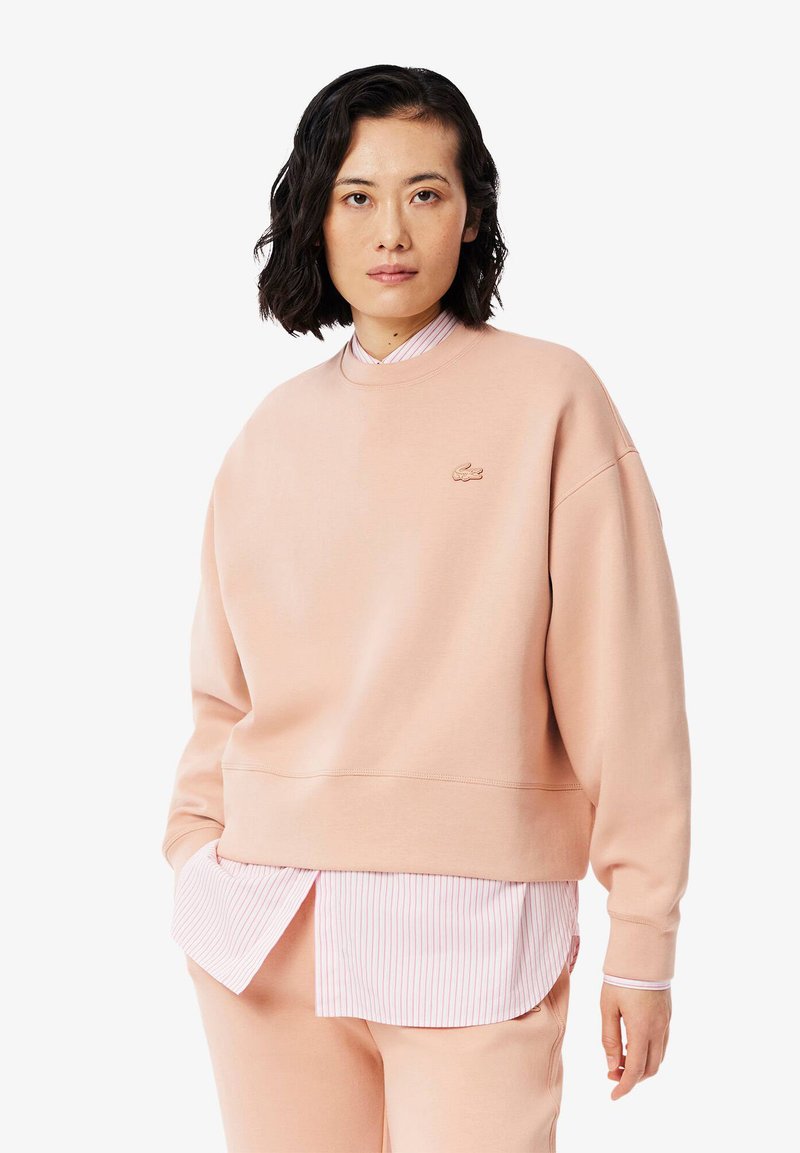 Rosa hettegenser laget av mykt materiale, har rund hals, ribbestrikkede mansjetter og en liten logo. Laget over en rosa og hvit stripet skjorte.