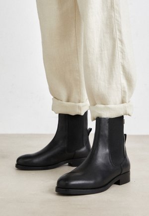 Sorte læder ankelstøvler med elastiske sidepaneler og en lav stakket hæl, parret med lyse beige opsmøgede bukser.