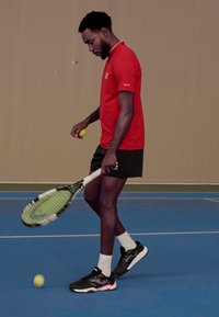 Tennista con una polo rossa e pantaloni corti neri, che tiene una racchetta e una pallina da tennis gialla, su un campo indoor blu.