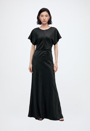 Femme debout portant une longue robe en satin noire avec des manches courtes et une fine ceinture nouée à la taille, sur un fond clair uni.