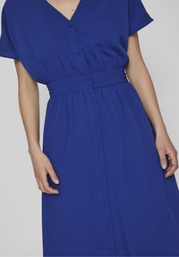 Modré šaty s krátkými rukávy, výstřihem do V a úzkým pasem. Obsahují plnou řadu knoflíků a splývavou sukni s jemnou texturou.