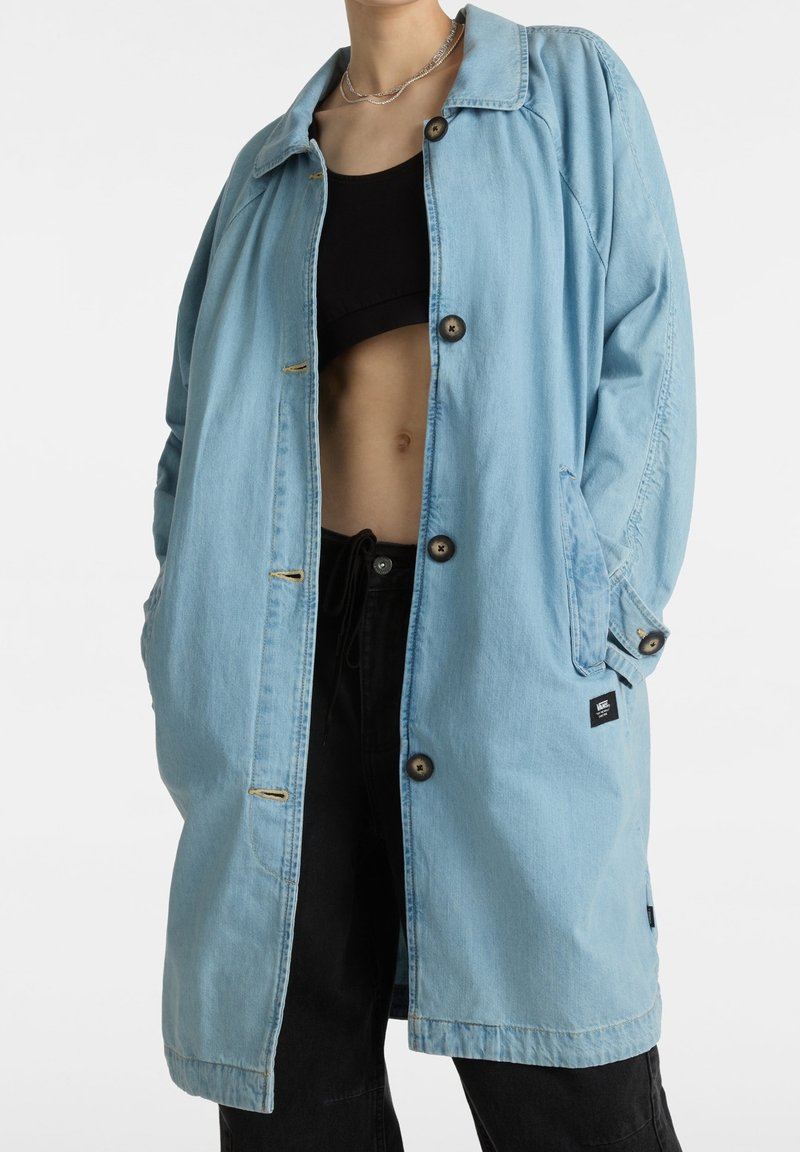 Lichtblauwe spijkerjas trenchcoat met lange mouwen, knoopsluiting, twee zijzakken en een zwart label aan de onderzijde links.