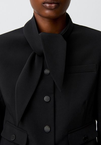 Persona che indossa un blazer nero con un grande dettaglio in tessuto annodato vicino al collo e tre bottoni visibili sul davanti.