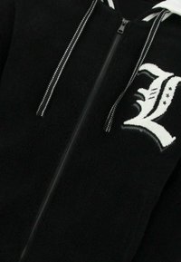Zwarte fleece jas met rits, met een wit letter "L" logo, contrasterende witte trekkoorden en een textuur in de stof.