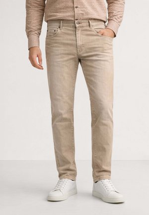 Uomo con jeans beige, camicia a maniche lunghe con motivo infilata dentro, scarpe da ginnastica bianche, in piedi con una mano in tasca.