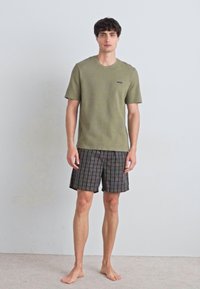 Khaki majica kratkih rukava s teksturiranim završetkom i logotipom, u kombinaciji s kratkim hlačama na kockice tamnijih tonova. Model stoji bos na neutralnoj pozadini.
