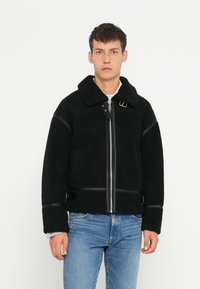 Schwarze Sherpa-Jacke mit Kragen, Reißverschluss und Lederakzenten. Verfügt über ein kastiges Design und elastische Bündchen. Wird über einem grauen Hemd getragen.