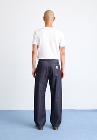 Dark blue denim pants with a loose fit, standard back pockets, and yellow stitching. Worn with a plain white t-shirt and boots.