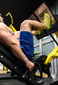 Beinpresse mit einer Metallplatte für die Füße; der Benutzer trägt knallgelbe Socken, einen schwarzen gepolsterten Sitz und blaue Shorts in einer Fitnessstudio-Umgebung.