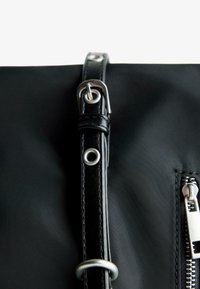 Black nylon bag featuring a leather strap with metal hardware, silver ring, and zipper detail on one side. Textured surface, sleek design.