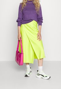 Fialový svetr, neonově zelená saténová sukni, růžová kabelka, bílé a černé tenisky, vrstvené textury, ležérní oblečení.