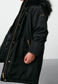 Parka noire matelassée avec capuche en fausse fourrure, fermetures éclair dorées et boutons-pression. Comprend des poches latérales et une taille cintrée pour une silhouette ajustée.