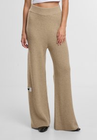 Beige pletené kalhoty s širokými nohavicemi a žebrovanou strukturou, vysokým pasem a malou logovou nášivkou na levé straně blízko lemu.