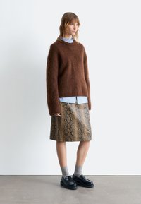 Brauner, flauschiger Pullover, darunter ein hellblaues kragenloses Hemd, ein gemusterter, sandfarbener Schlangenprint-Rock, graue Socken und schwarze Plateauschuhe.