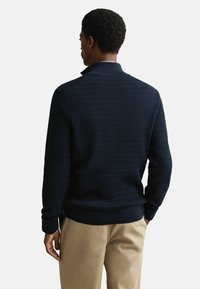 Marine gebreide trui met een gestructureerd patroon, staande kraag en geribde manchetten en zoom. Gecombineerd met beige broeken.