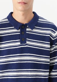 Marineblauwe en witte gestreepte polo met een gebreide structuur, omgeslagen kraag en drie zwarte knopen bij de knoopsluiting.