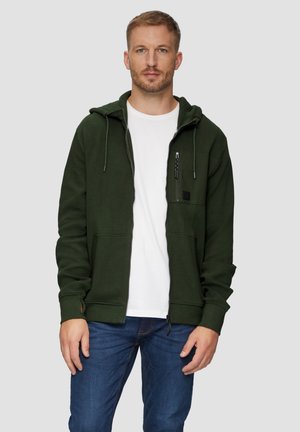 Haut zippé vert avec poches avant et capuche à cordon. Porté par-dessus un t-shirt blanc, associé à un jean bleu. Présente une poche poitrine zippée.