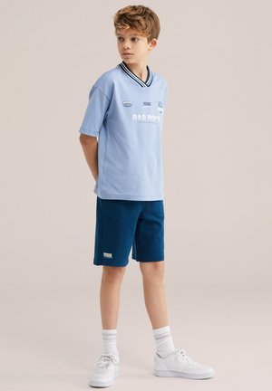 Băiat stând cu mâinile la spate, purtând un tricou grafic albastru deschis, pantaloni scurți bleumarin, șosete albe și adidași albi, pe un fundal neutru.