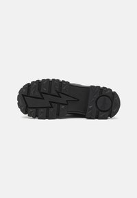 Suola di scarpa in gomma nera con profonde e frastagliate scanalature, caratterizzata da un design a fulmine e un dettaglio con logo circolare. Superficie liscia e texture.