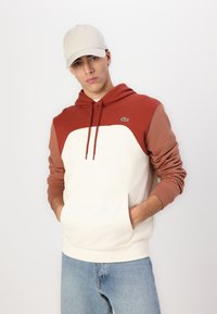 Felpa con cappuccio color crema e ruggine con maniche lunghe; presenta una tasca frontale e un logo ricamato sul petto. Accompagnata da un cappello beige.
