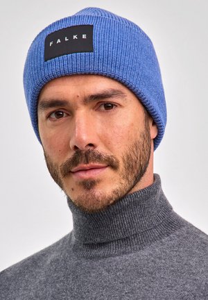 Man met een blauwe gebreide beanie met het label "FALKE" en een grijze coltrui, op een neutrale achtergrond.