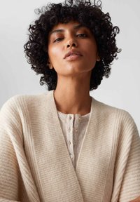 Beige Strickjacke mit gerippter Textur, V-Ausschnitt und Knöpfen entlang der Vorderseite. Das Model hat lockiges Haar und einen natürlichen Hautton.