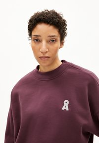 Burgunderfarbenes Sweatshirt aus weichem Material mit hohem Kragen und einem weißen gestickten Logo auf der linken Brust.