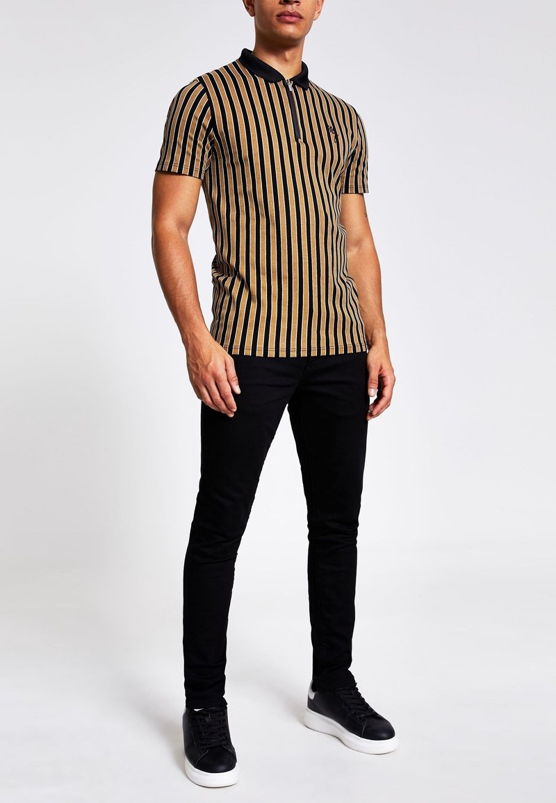 Homme portant un polo à manches courtes avec des rayures verticales noires et beige, un pantalon noir et des baskets noires à semelles blanches.