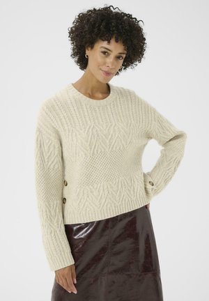 Pull en maille de couleur beige clair avec des motifs texturés, des poignets côtelés et des boutons décoratifs sur le côté. Assorti à une jupe bordeaux brillante.