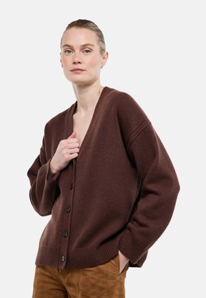 Femme aux cheveux plaqués en arrière, portant un cardigan marron boutonné et un pantalon beige, posant avec une main dans la poche et l'autre tenant le cardigan.