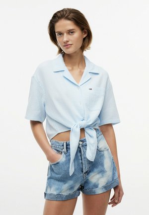 Giovane donna che indossa una camicia azzurro chiaro a maniche corte annodata in vita e shorts in denim scolorito a vita alta.