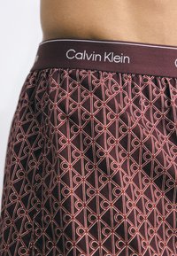 Calvin Klein bokseršorti tumši sarkanā audumā ar ģeometrisku rakstu, kas sastāv no savstarpēji savienotiem līdzīgiem formām, un pretrunīgu logotipa jostu.