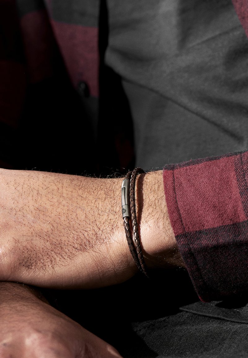 Brun flätad läderarmband med metallspänne, virad runt en handled, vilande på en texturerad yta. Mörkrutig skjorta synlig.