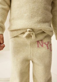Beige knitted sweater set with a relaxed fit. Features a tie at the waist and pink "NYC" lettering on pants. Soft, textured fabric.