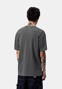 Uomo con capelli neri che indossa una maglietta grigia scura larga e pantaloni in denim scuri, che mostra tatuaggi su collo e braccia, in piedi di spalle.