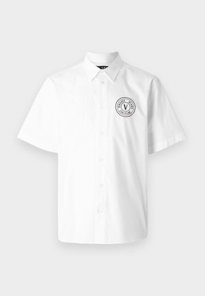 Camisa branca de mangas curtas em algodão, com colarinho abotoado e um logotipo preto circular na área do lado esquerdo do peito.