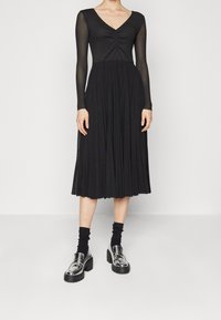 Černé dlouhé šaty s výstřihem do V, s texturovaným živůtkem a skládací sukní. V kombinaci s lesklými stříbrnými mokasíny a černými ponožkami.