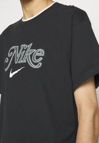 Svart bomullströja med en stor broderad "Nike"-logotyp i vitt och grått samt en liten vit swoosh, rund hals och korta ärmar.