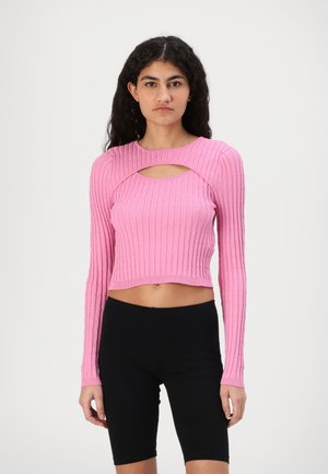 Maglione rosa a coste, corto, con maniche lunghe e dettaglio con ritaglio frontale. Abbinato a pantaloni ciclisti neri.