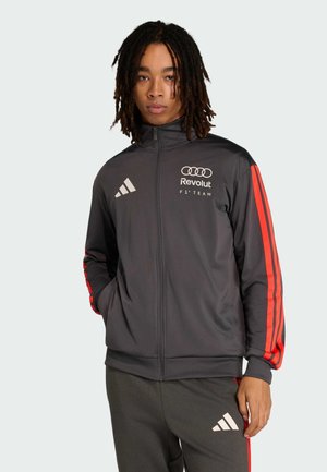 Mann mit schulterlangen Dreadlocks, der eine schwarze Adidas-Jacke mit roten Streifen und dem Revolut F1 Team-Logo trägt, steht vor hellem Hintergrund.