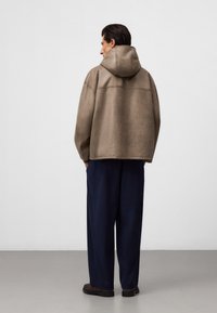 Brązowa, oversized bluza z kapturem o teksturowanym wykończeniu, z opadającymi ramionami i kontrastowym przeszyciem, w połączeniu z luźnymi granatowymi spodniami.